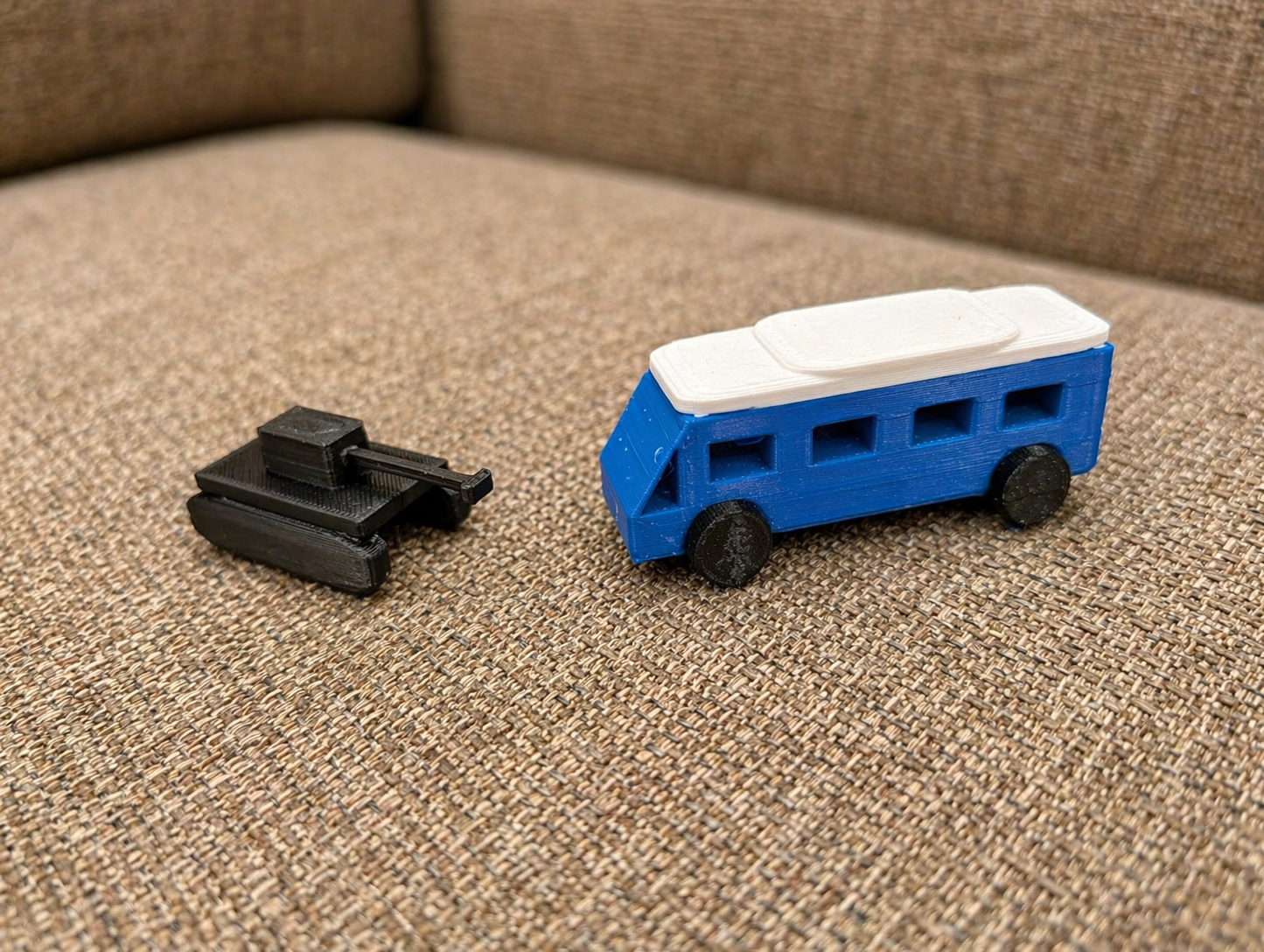 Two small 3D printed vehicles, one black tracked vehicle and one blue camper van with a white roof, displayed on a couch.